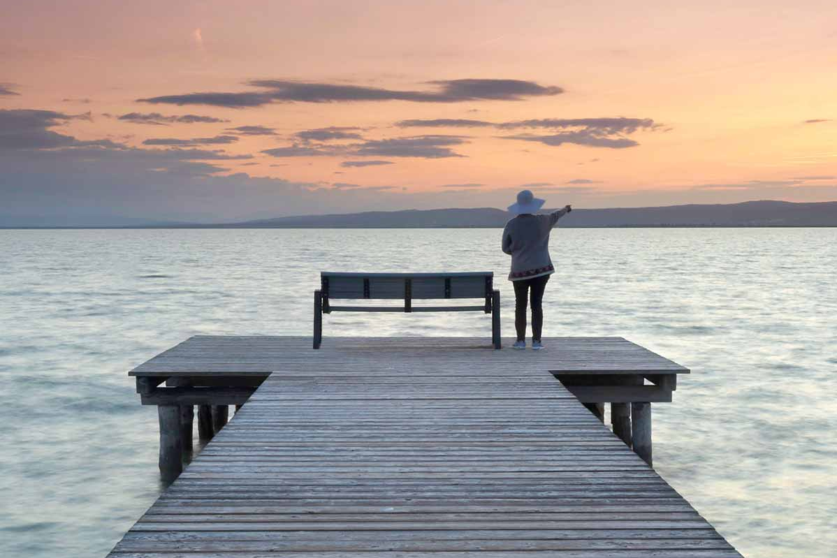 Eine Frau mit Hut steht auf einem Steg im See und zeigt auf eine Hügelkette